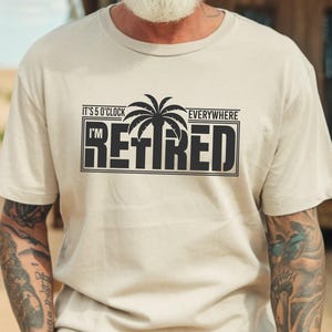 May include: Beige t-shirt with the text "It's 5 O'Clock Everywhere I'm Retired" in a black rectangular design with a palm tree graphic. The shirt is worn by a person wearing a hat and sunglasses.