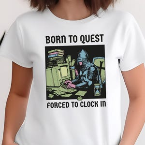 May include: White t-shirt with a graphic of a knight at a computer, with the text "BORN TO QUEST" above and "FORCED TO CLOCK IN" below. The knight is wearing armor and typing. The shirt is a crew neck.