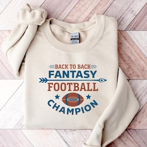 May include: A beige sweatshirt with the text "Back to Back Fantasy Football Champion" in brown, blue, and white lettering. A football graphic is centered below the text.