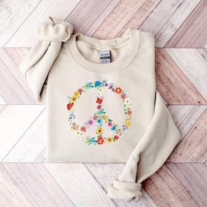 May include: A beige crewneck sweatshirt featuring a central graphic of a peace sign composed of various colorful flowers including red, yellow, blue, and white blossoms. The garment has long sleeves with ribbed cuffs and a Gildan brand label at the neck.