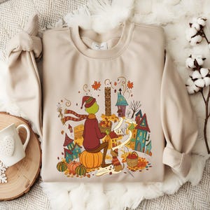 May include: A beige crewneck sweatshirt featuring a colorful graphic of a green character wearing a red scarf and hat, sitting on a pumpkin in a village setting with autumn leaves, small houses, and a dog, all rendered in a detailed illustration.