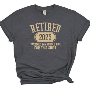 May include: Dark gray t-shirt with rolled sleeves. The shirt features the text "RETIRED 2025" in a beige font, with the phrase "I WORKED MY WHOLE LIFE FOR THIS SHIRT" below. The shirt is a casual wear item.