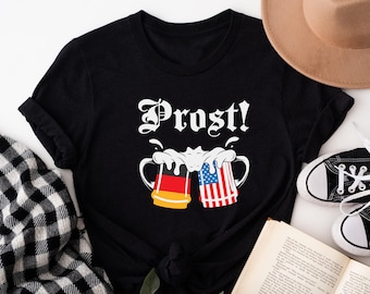 Könnte beinhalten: Schwarzes T-Shirt mit einer weißen Grafik von zwei Bierkrügen, die anstoßen. Die Krüge sind mit der deutschen und amerikanischen Flagge verziert und der Text "Prost!" befindet sich darüber.