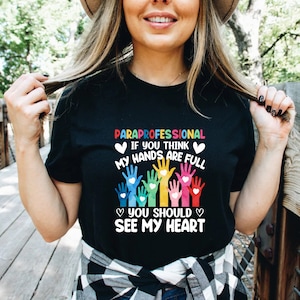 May include: Black t-shirt with the text "PARAPROFESSIONAL" and "IF YOU THINK MY HANDS ARE FULL YOU SHOULD SEE MY HEART". The text is surrounded by colorful handprints with hearts.