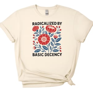 May include: Cream-colored t-shirt featuring a floral design with red and blue flowers and leaves. The text "RADICALIZED BY BASIC DECENCY" is printed above and below the floral graphic. The shirt has short sleeves and a crew neck.