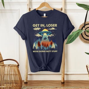 May include: A navy blue short-sleeve t-shirt featuring a graphic print of a UFO hovering over a forest with mountains in the background. The text "GET IN, LOSER" is printed above the UFO, and "WE'RE DOING BUTT STUFF" is printed below the forest.