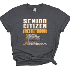 May include: Dark gray t-shirt with the words "SENIOR CITIZEN TEXTING CODE" in white and yellow lettering. Below the text is a list of texting acronyms and their meanings, such as "BFF" for "Best Friend Fell".