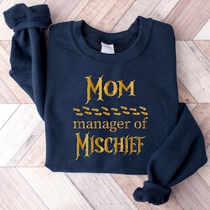 May include: A navy blue long-sleeve crewneck sweatshirt featuring gold-colored text that reads "MOM manager of MISCHIEF" with a horizontal row of small gold-colored footprints positioned between the words "MOM" and "manager of".