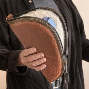 Op de afbeelding: Een bruine leren etui met een ritssluiting, gevoerd met witte vacht. De etui heeft een donkergroene rand en een metalen ritstrekker. Een blauw object is zichtbaar in de etui. De etui wordt opengehouden en toont de inhoud.