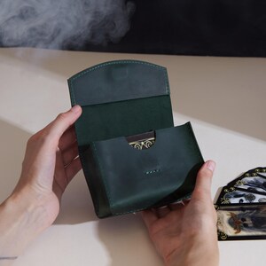 Puede incluir: Una caja para cartas de cuero verde oscuro con cierre de solapa y costuras visibles. La caja está abierta, revelando una parte de una baraja con patrones dorados y negros en los bordes, mientras que otras cartas están extendidas al lado.