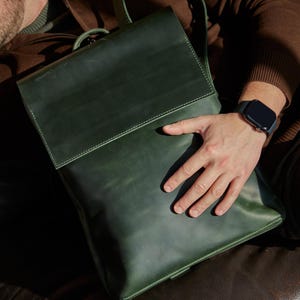 Könnte beinhalten: Ein Rucksack aus waldgrünem Leder mit einem großen, rechteckigen Klappverschluss und sichtbaren Nähten an den Kanten. Die Tasche hat eine glatte, matte Oberfläche und verfügt über einen Tragegriff sowie Schultergurte in einem klaren Design.