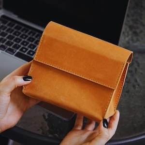 Può includere: Una custodia rettangolare in pelle color cuoio con chiusura a patta ripiegabile e cuciture visibili lungo i bordi. La custodia presenta una texture liscia e opaca e un design strutturato, tenuta in modo da mostrare chiaramente il profilo frontale e laterale.