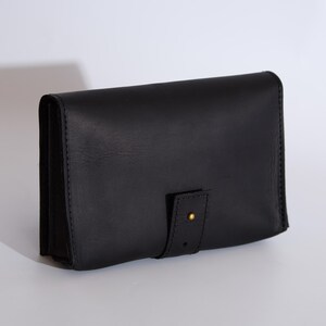May include: A rectangular black leather wallet featuring a smooth finish and visible stitching along the edges. The wallet includes a front flap secured by a vertical leather tab with a small, circular brass-colored metal stud fastener.