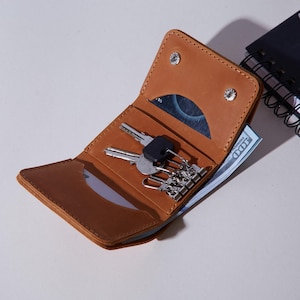 May include: A tan leather key holder wallet featuring a fold-over design with two silver-tone snap closures. The interior contains a metal key ring mechanism with multiple clips, a dedicated card slot, and a compartment holding a 100 dollar bill.