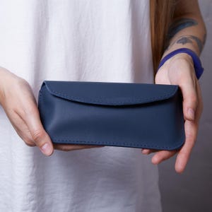 May include: A rectangular navy blue leather case with a curved flap closure and visible stitching along the edges. The case features a smooth matte finish and a structured design, providing a secure enclosure for small items like glasses or pens.