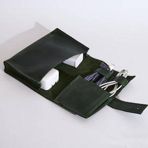 Op de afbeelding: Een donkergroene lederen organizer-etui met meerdere binnenvakken. Het etui is geopend en bevat twee witte stroomadapters en twee opgerolde oplaadkabels die worden vastgehouden door lederen lussen, met een riemsluiting aan de rechterkant.