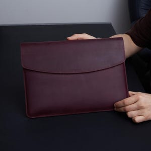 Peut inclure: Une housse pour ordinateur portable en cuir bordeaux profond, dotée d'une finition mate lisse et d'un rabat incurvé. L'étui rectangulaire présente des bords cousus nets et un design épuré, offrant une protection sécurisée pour un ordinateur.
