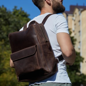 Può includere: Uno zaino in pelle marrone scuro caratterizzato da un ampio scomparto principale con chiusura a zip e una tasca frontale più piccola con patta. Lo zaino è dotato di spallacci regolabili e una finitura liscia e opaca, indossato sopra una t-shirt bianca.