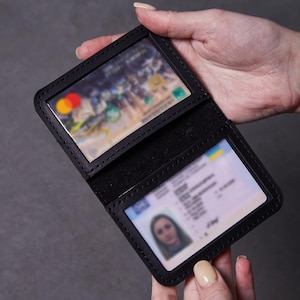 Peut inclure: Un portefeuille noir à deux volets doté de deux fenêtres en plastique transparent pour afficher des cartes d'identité ou des cartes de crédit. Le portefeuille est construit avec des coutures noires visibles le long des bords.