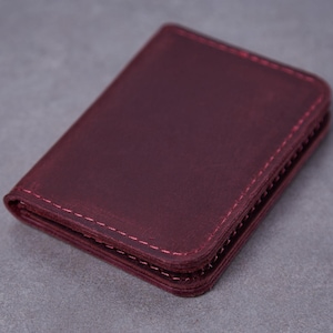 May include: A rectangular burgundy leather card holder featuring rounded corners and matching burgundy stitching along the edges. The wallet has a smooth, matte finish and a folded construction with visible layers of leather stacked on the side.
