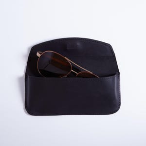 Könnte beinhalten: Ein schwarzes Brillenetui aus Leder mit geschwungenem Klappverschluss, das eine goldfarbene Fliegerbrille mit dunkelbraunen Gläsern enthält. Das Etui weist sichtbare Nähte an den Rändern und eine glatte, matte Oberfläche auf der Außenseite auf.