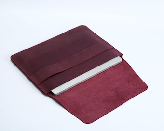 May include: A deep burgundy leather laptop sleeve with a fold-over flap closure. The sleeve features a horizontal strap across the front panel and contains a thin silver laptop partially inserted into the main compartment.