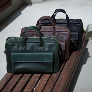 Pode incluir: Três pastas retangulares de couro dispostas em fila, com um bolso frontal com aba, duas alças superiores e um compartimento principal com zíper. As bolsas são verde-escuras, castanho-escuras e pretas, com um acabamento fosco e liso.