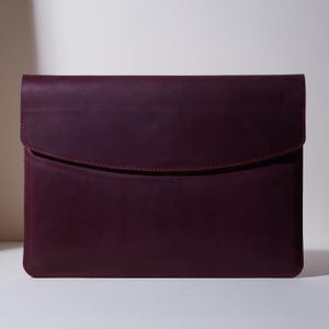May include: A rectangular dark burgundy leather laptop sleeve featuring a smooth matte finish, a curved flap closure, and tonal stitching along the edges. The sleeve has a clean, structured design with rounded corners and a flat, minimalist profile.