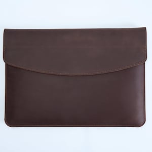 Pode incluir: Uma capa para portátil em couro castanho escuro com um acabamento mate suave e um fecho de aba curva estilo envelope. O estojo retangular tem bordas costuradas limpas e um design simples, proporcionando uma cobertura protetora para um portátil.