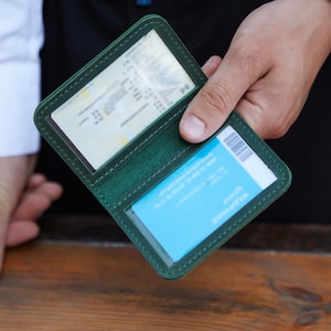 May include: A green leather card holder, open to display a driver's license and a blue card. The holder is held in a person's hand, with a wooden surface in the background. The holder has a clear plastic window.
