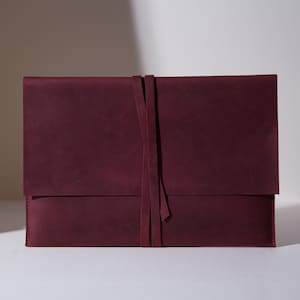 Könnte beinhalten: Eine tiefburgunderrote Dokumentenmappe aus Leder im rechteckigen Umschlag-Design mit glatter, matter Oberfläche. Die Mappe wird durch zwei lange, schmale Lederriemen verschlossen, die vertikal um die Mitte gewickelt sind und die Klappe halten.