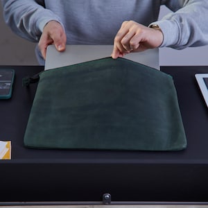 Könnte beinhalten: Eine dunkelgrüne Laptoptasche mit Reißverschluss. Die Tasche besteht aus einem matten, glatten Material und hat ein schmales, rechteckiges Profil, das dazu dient, einen dünnen silbernen Laptop sicher im Hauptfach zu halten.