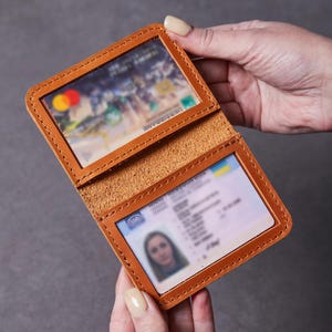 Könnte beinhalten: Ein beigefarbener Kartenhalter aus Leder mit zwei durchsichtigen Kunststofffenstern für Ausweise oder Kreditkarten. Das Etui ist mit sichtbaren Nähten an den Kanten versehen und lässt sich aufklappen, um zwei Karten zu zeigen.