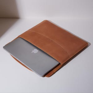 Könnte beinhalten: Eine hellbraune Laptoptasche aus Leder mit glatter, matter Oberfläche, die teilweise einen silbernen Laptop mit einem sichtbaren weißen Apfel-Logo auf dem Deckel umschließt. Die Tasche hat ein schlichtes Design mit sichtbaren Nähten an den Kanten.