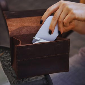 Op de afbeelding: Een donkerbruine lederen opbergdoos met een klepdeksel, met daarin een wit elektronisch apparaat en een witte oplaadkabel. De doos heeft zichtbare stiksels langs de randen en een matte afwerking, wat een gestructureerde houder biedt voor accessoires.