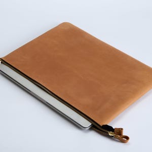 Pode incluir: Uma capa de couro castanho para portátil com um acabamento mate suave e um fecho de correr em metal escuro. A capa é retangular com cantos arredondados e contém um portátil prateado parcialmente visível.