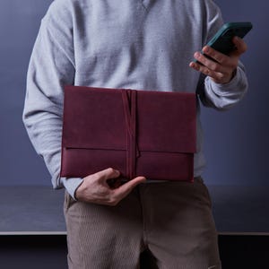 Peut inclure: Une pochette à documents en cuir bordeaux profond présentant un design rectangulaire de style enveloppe avec une longue et fine lanière en cuir qui s'enroule verticalement autour du centre pour sécuriser la fermeture à rabat. La surface est mate.