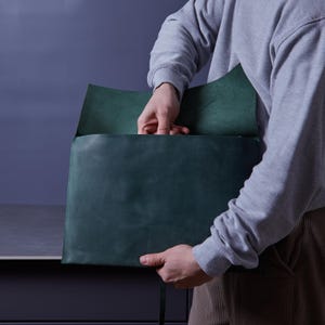 May include: A dark green leather document folder with a flap closure. The folder features a smooth, matte finish and visible stitching along the edges. The rectangular design is held open, revealing the interior compartment and the texture of the leather.