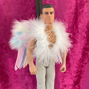 Puede incluir: Una muñeca con un traje de plumas blancas con un ala de plumas de arcoíris. La muñeca tiene una cara esculpida con cabello castaño y ojos azules.