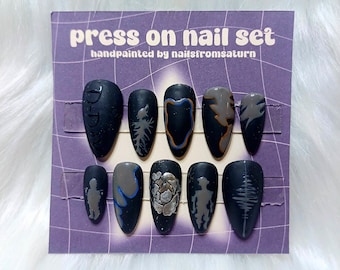 May include: A set of ten black press-on nails with different designs. The designs include a pine tree, a flower, a silhouette of a person, a wave, and a geometric shape. The nails are hand-painted and have a matte finish. The card reads "press on nail set" and "handpainted by nailsfromsaturn".