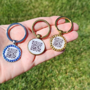 May include: Three keychains with QR codes are displayed against a green background. The keychains have different metal finishes: silver, bronze, and gold. The silver keychain has blue rhinestones, the gold keychain has gold rhinestones, and the bronze keychain is plain.