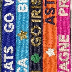 Puede incluir: Cinco cinturones de cuentas con diferentes nombres y logotipos de equipos. Los cinturones son azules, azul claro, azul marino, naranja y rosa. Los nombres de los equipos son Wildcats, ULGA, Irish, Astros y Champagne.