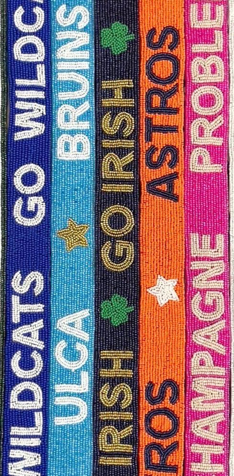 Puede incluir: Cinco cinturones de cuentas con diferentes nombres y logotipos de equipos. Los cinturones son azules, azul claro, azul marino, naranja y rosa. Los nombres de los equipos son Wildcats, ULGA, Irish, Astros y Champagne.