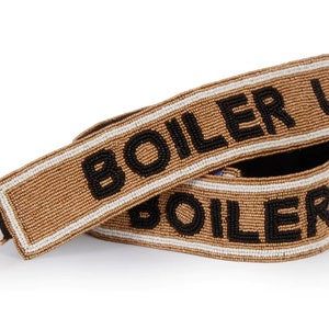 May include: A brown beaded bag strap with the text "BOILER UP" in black beads. The strap is folded in half and the ends are visible.