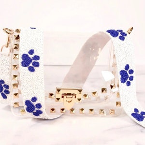 May include: Clear plastic crossbody bag with a white beaded strap featuring blue paw print designs. The bag has gold studs and a gold clasp.