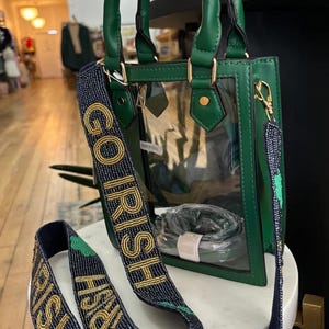 Puede incluir: Un bolso de mano verde y transparente con una correa de cuentas doradas que dice "Go Irish".