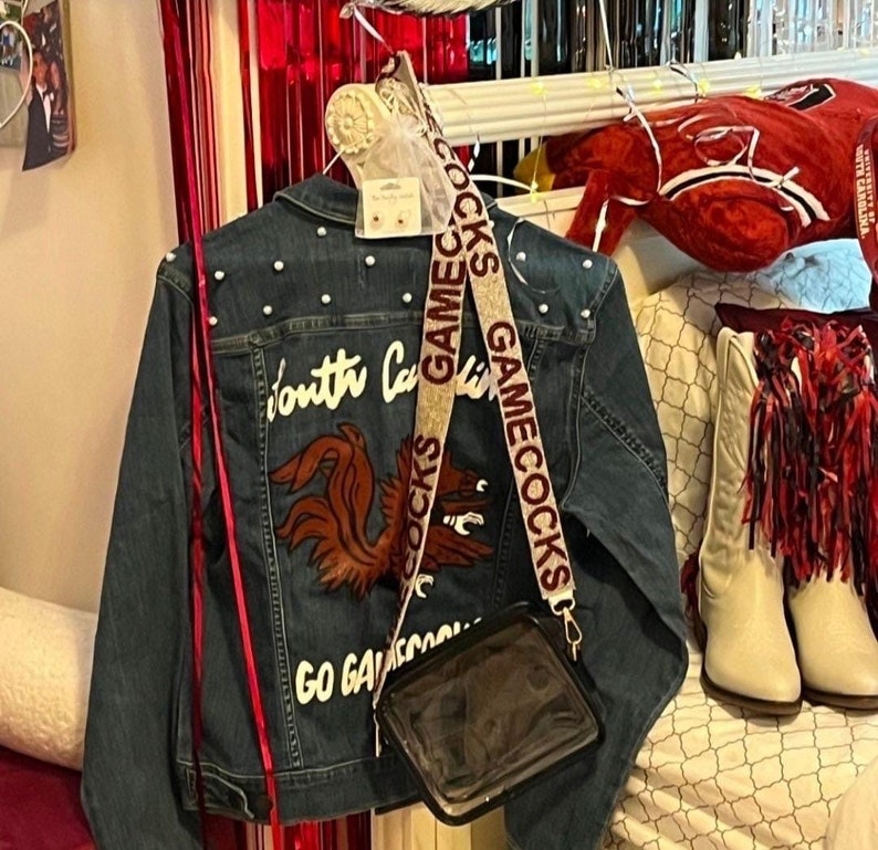 May include: A blue denim jacket with pearl embellishments and a red and white embroidered design that says "South Carolina Gamecocks Go Gamecocks". The jacket has a clear plastic bag attached to it with a beige strap that says "Gamecocks Gamecocks".