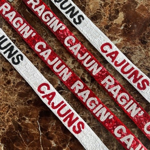 May include: Four white and red sequined fabric strips with the text "CAJUNS" and "RAGIN' CAJUN" in black lettering.