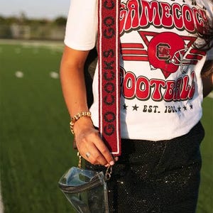 May include: A white t-shirt with a red and white graphic that says "CAROLINA GAMECOCK FOOTBALL EST. 1883". The shirt is being worn with a black sequined skirt and a clear crossbody bag with a red and white beaded strap that says "COCKS GO COCKS".