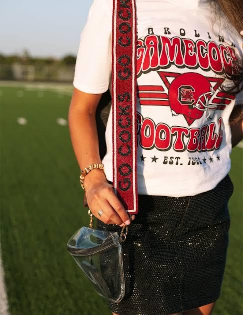 May include: A white t-shirt with a red and white graphic that says "CAROLINA GAMECOCK FOOTBALL EST. 1883". The shirt is being worn with a black sequined skirt and a clear crossbody bag with a red and white beaded strap that says "COCKS GO COCKS".